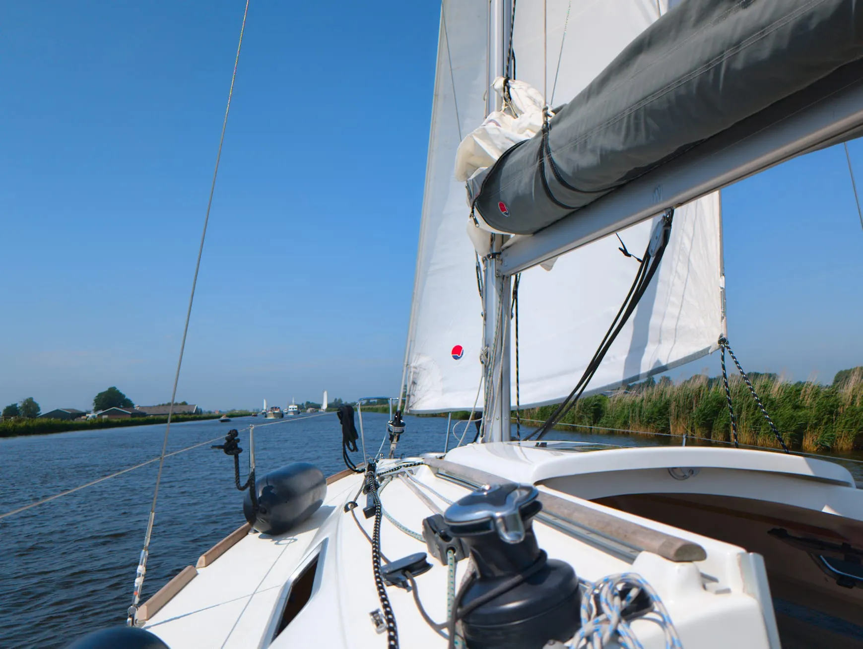 View from a sailing boat beneath white sails, with blue sky and a reed-lined waterway.